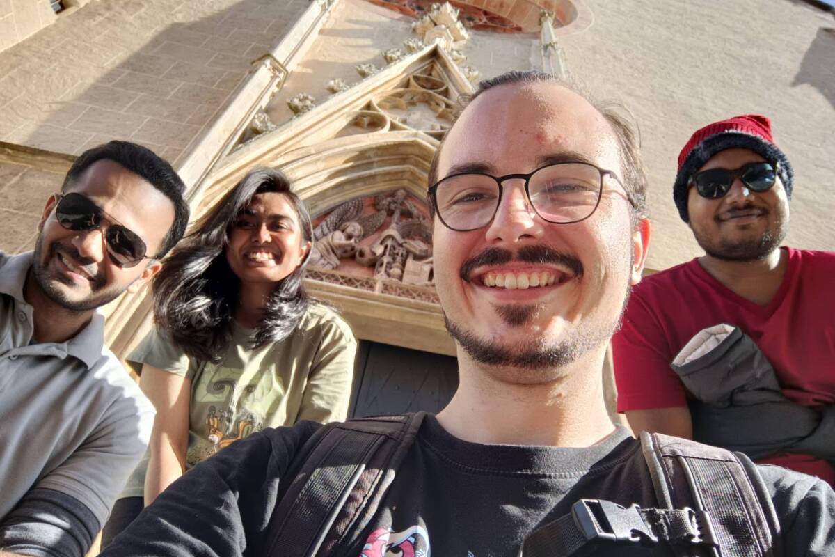 Das Bild zeigt vier Teilnehmer:innen des Welcome Buddy Programms: zwei Buddies und zwei Mentees. Sie haben sich bei einer gemeinsamen Aktivität vor einer Kirche platziert und ein Selfie gemacht.