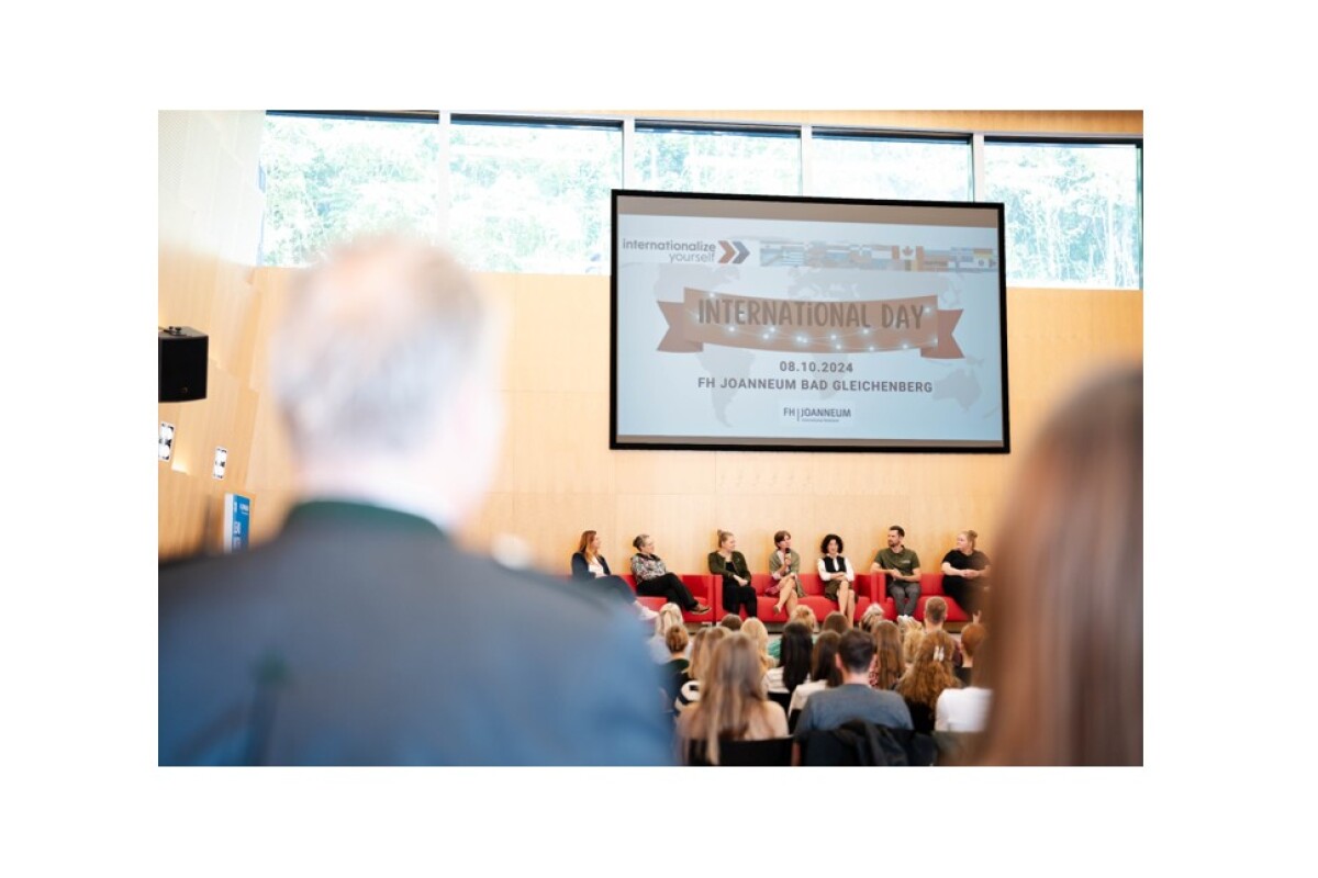 Eine Bühne mit Menschen, die auf Sofas sitzen, ist zu sehen. Eine Dame hält ein Mikrofon in der Hand und erzählt. Vor der Bühne erkennt man Publikum.