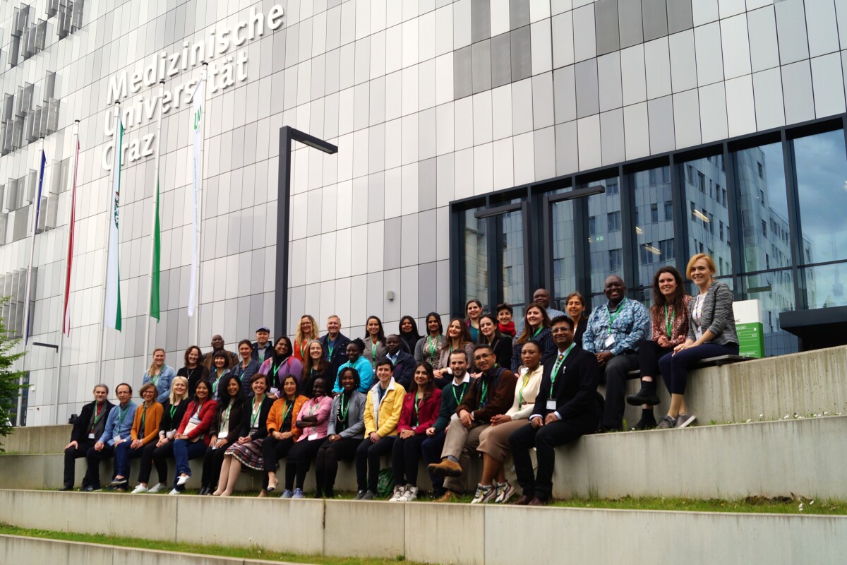 The group of international participants sits in front of the Med Uni Graz building. The Med Uni Graz lettering and some flags can be seen in the background.
