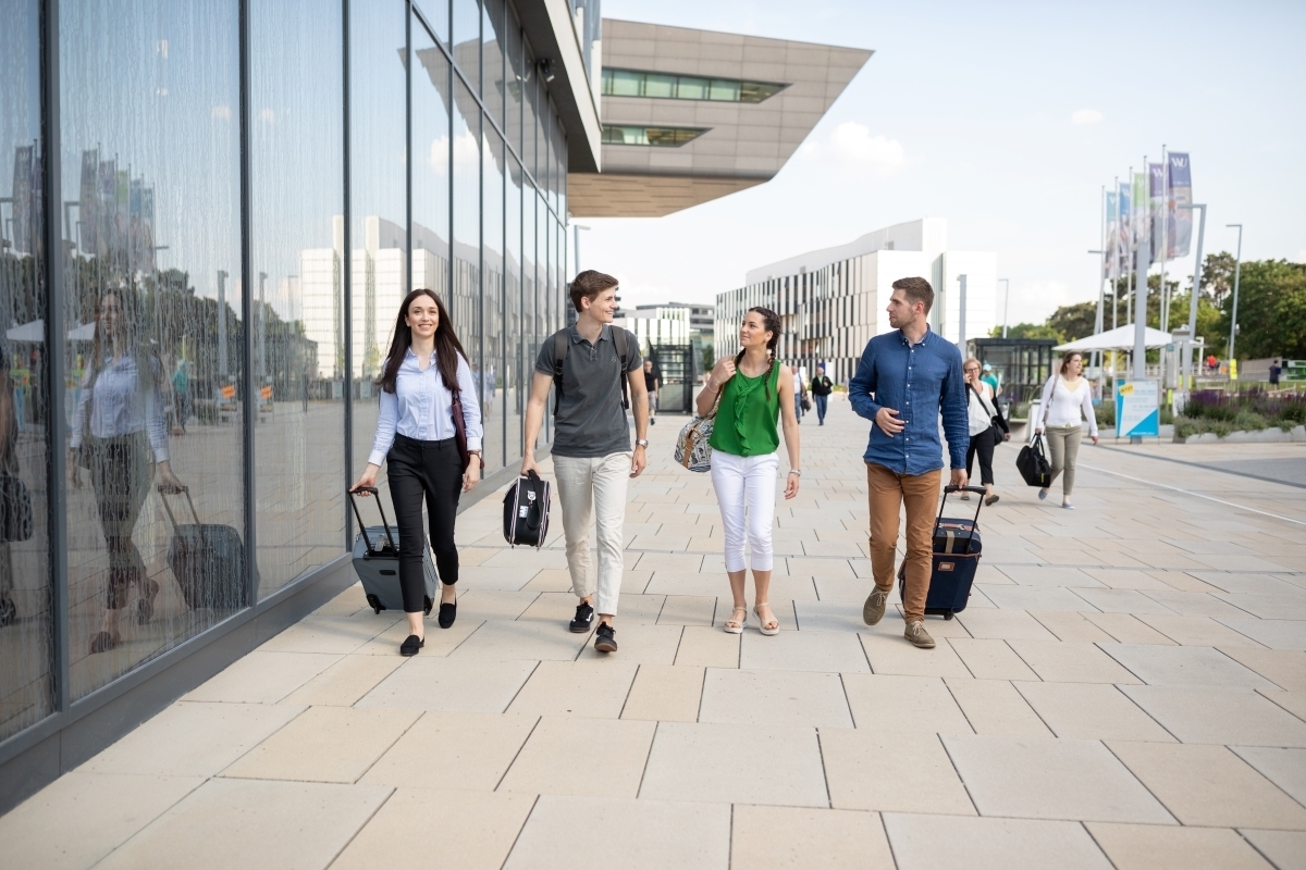Darstellung von WU-Studierenden. Die Studierenden bewegen sich mit Trolleys über den Campus, um die Wichtigkeit internationaler Orientierung - Global Mindset - im Studium zum Ausdruck zu bringen.